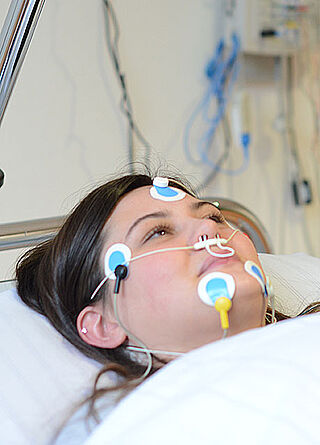 Eine Patientin liegt mit verschiedenen Elektroden im Gesicht und einer Sauerstoffbrille im Bett.